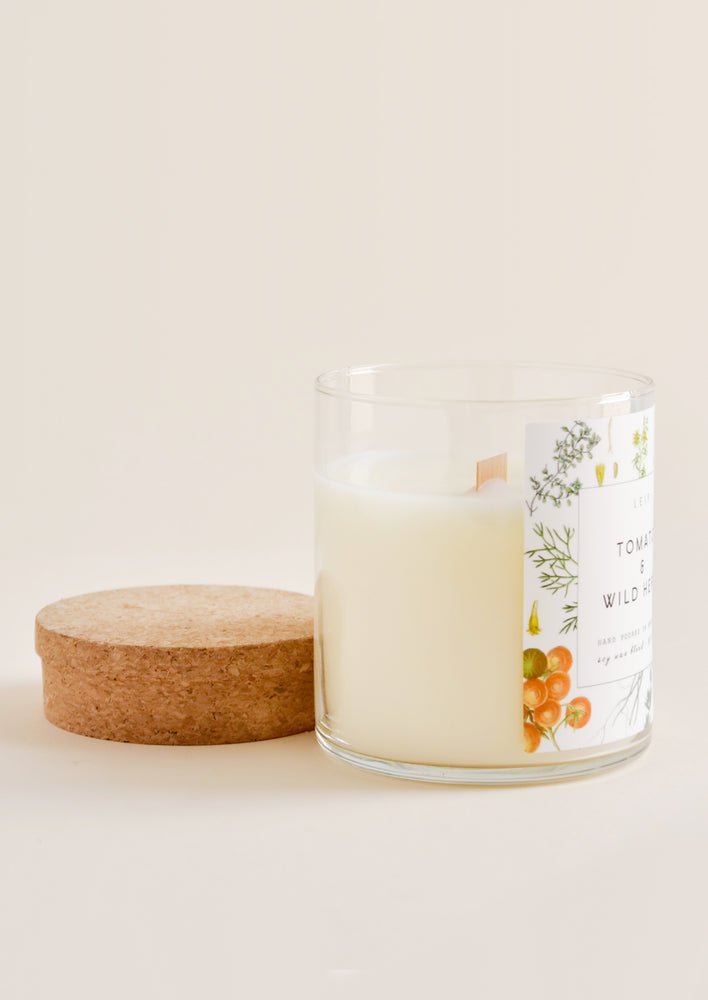A glass candle with botanical label and wooden wick sits next to its cork lid.
