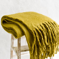 1: A chartreuse mohair blanket with long tassel fringe, folded on a stool.