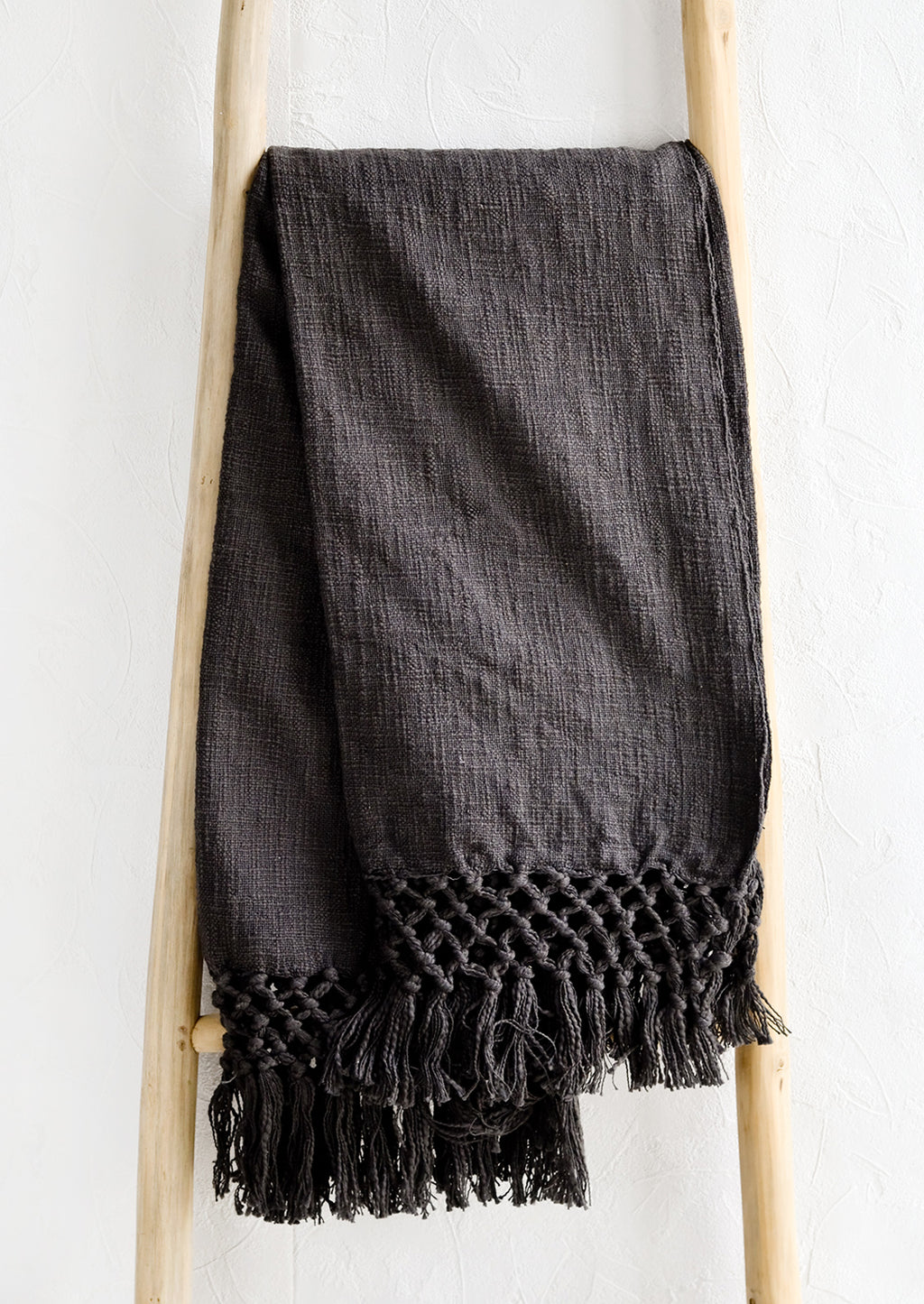 Carbon: A dark grey cotton throw blanket with knotted open weave trim, displayed on a ladder.