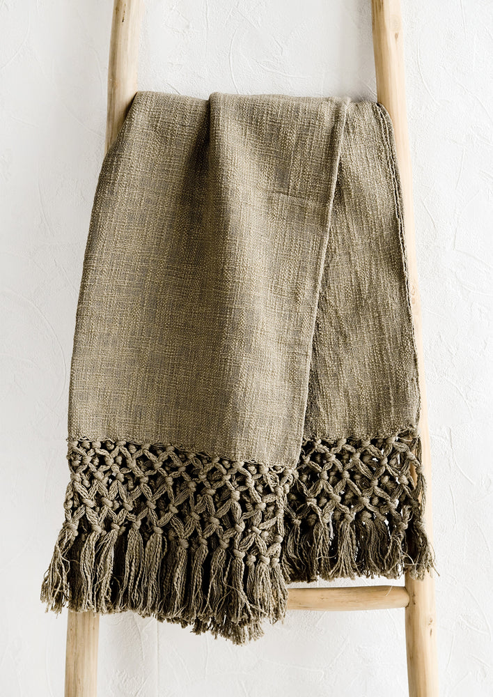 Fatigue: An army green cotton throw blanket with knotted open weave trim, displayed on a ladder.