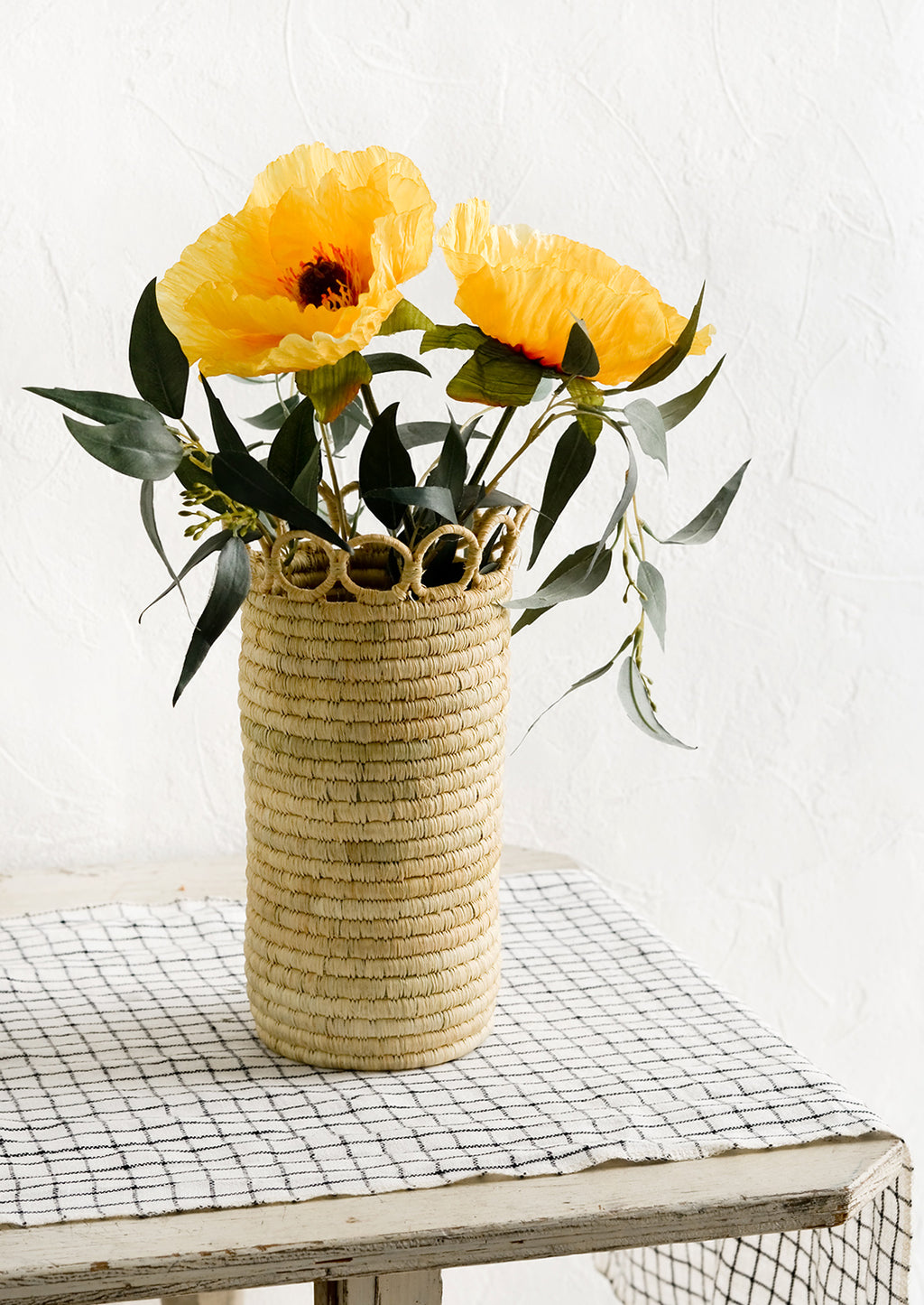 2: A cylindrical raffia vase on a linen-clad table with eucalyptus and poppies.