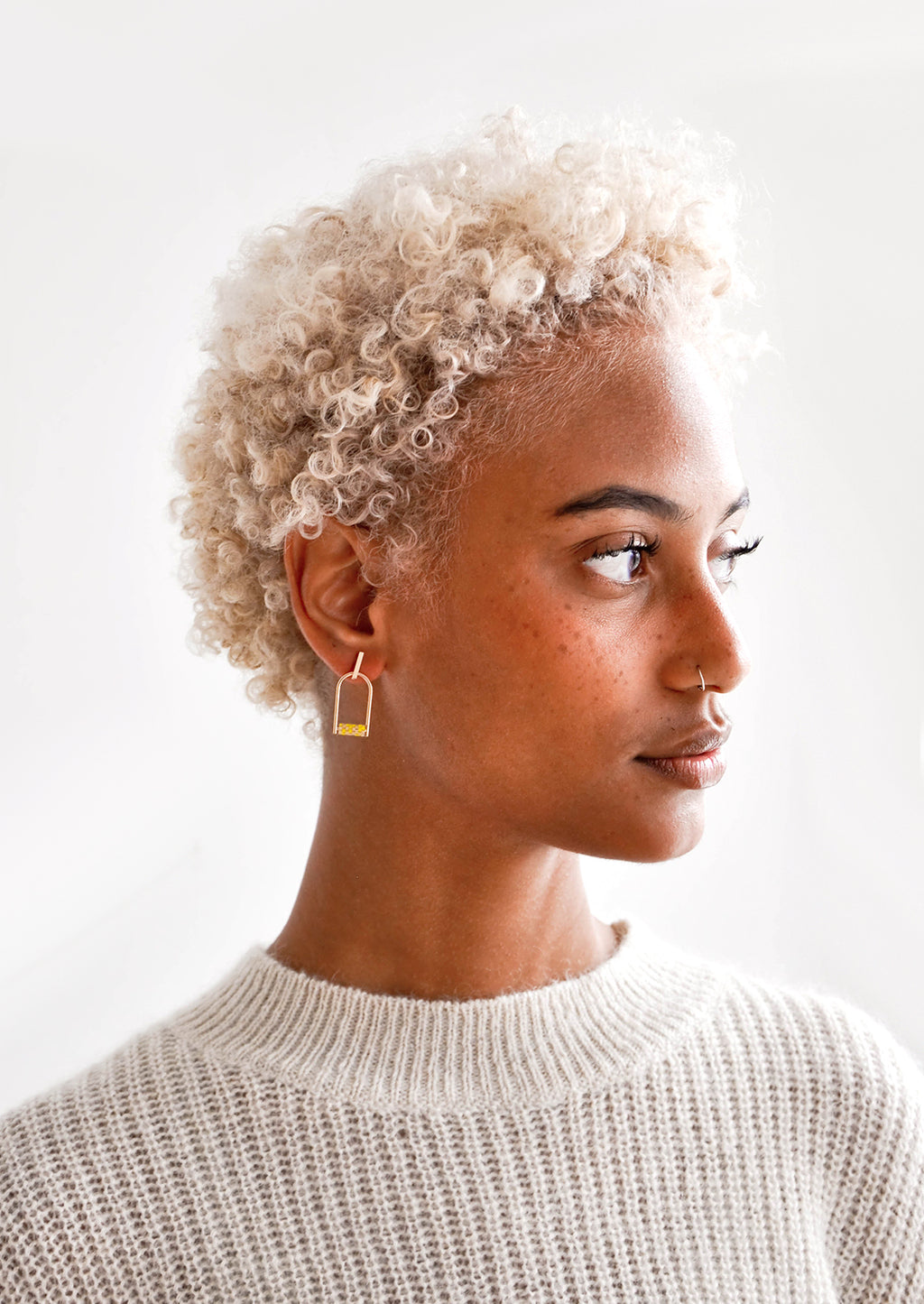 3: Model wears arced gold earrings with yellow beads and a cream colored sweater.