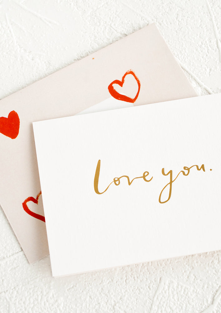 A white greeting card with golden cursive reading "Love you", paired with heart print envelope.