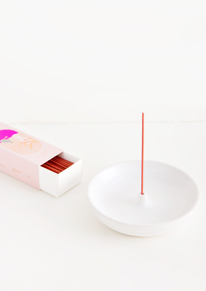 A simple white curved ceramic incense holder with a pink stick of incense and a pink box of incense next to it.