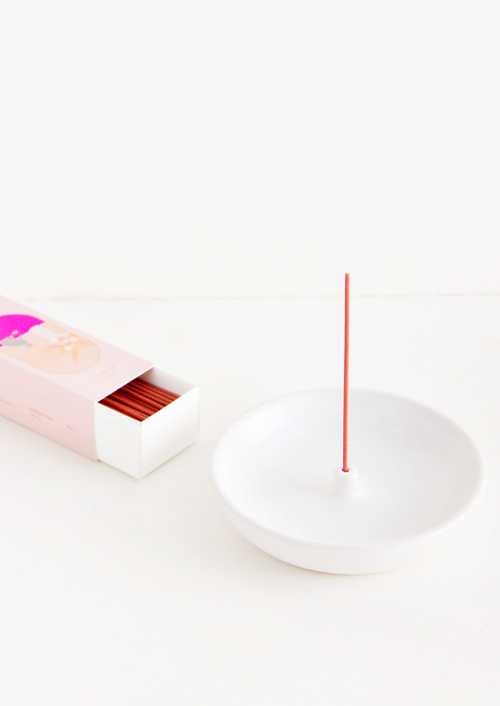 1: A simple white curved ceramic incense holder with a pink stick of incense and a pink box of incense next to it.