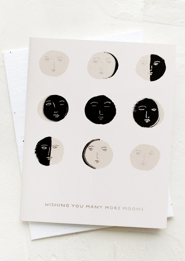 Greeting card with illustrated moon faces and silver text reading "Wishing you many more moons"