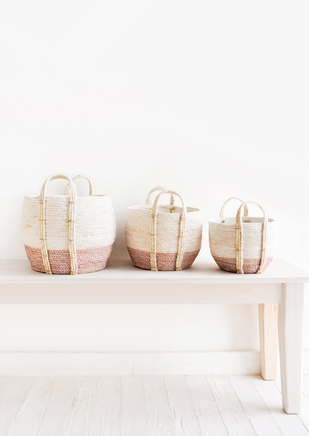 Dusty Rose / Low [Small]: Line up of three round storage baskets in incremental sizes, handles attached at sides, band of contrasting pink color along bottom.