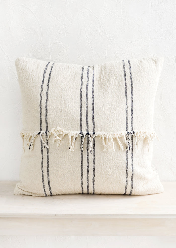 A natural cotton throw pillow with vertical dark stripes and fringed panel at center.