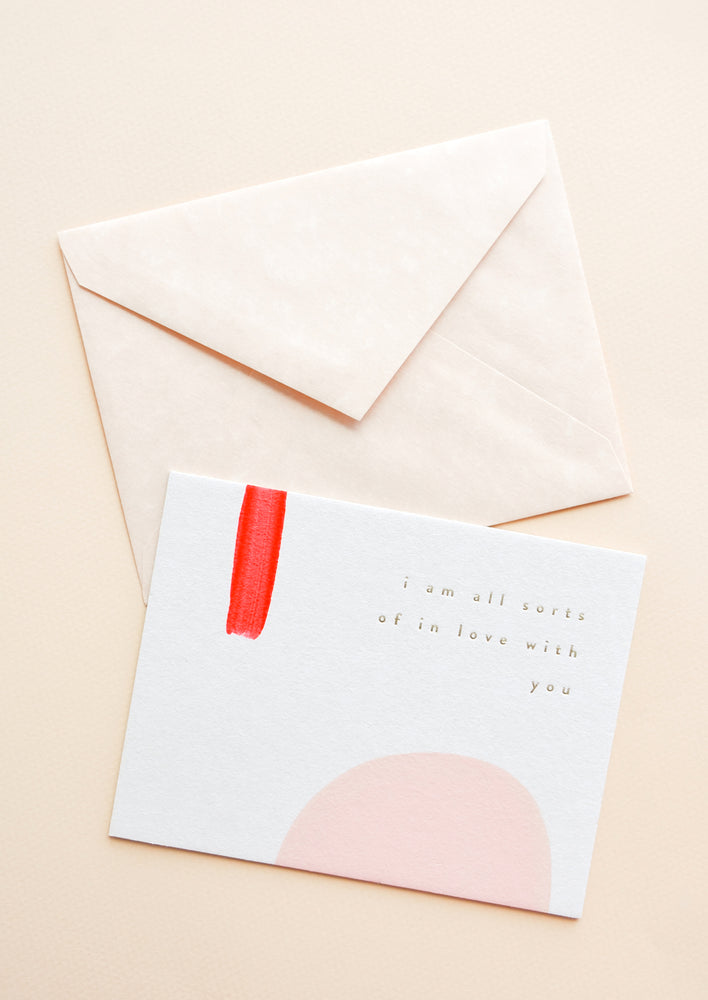 1: A pale tan envelope and white greeting card with a rectangle of red paint, a semicircle of pink paint, and gold foil text reading "I'm all sorts of in love with you."