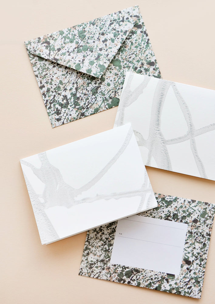 1: Two envelopes patterned with green and gray paint splatter alongside two white greeting cards with thin strokes of gray paint.