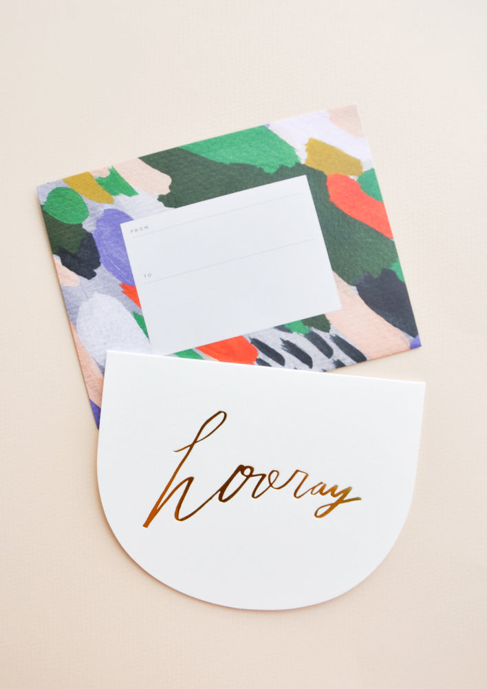 A colorful patterned rectangular envelope with an accompanying greeting card in a white semicircle with gold foil script reading "hooray."