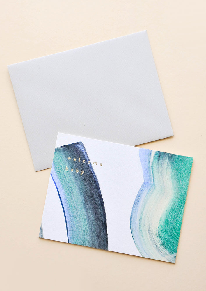 2: A grayish envelope and a white greeting card with multicolored paint strokes in blue, green, beige, and black and the words "welcome baby" in small lowercase gold foil letters at the top left.