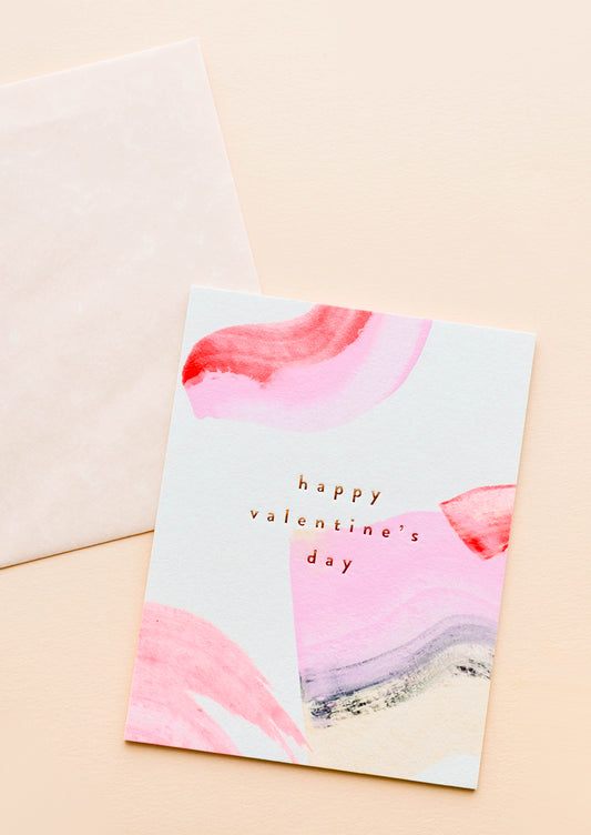 A beige envelope and white greeting card patterned with pink and red paint strokes reading "happy valentine's day" in gold foil.