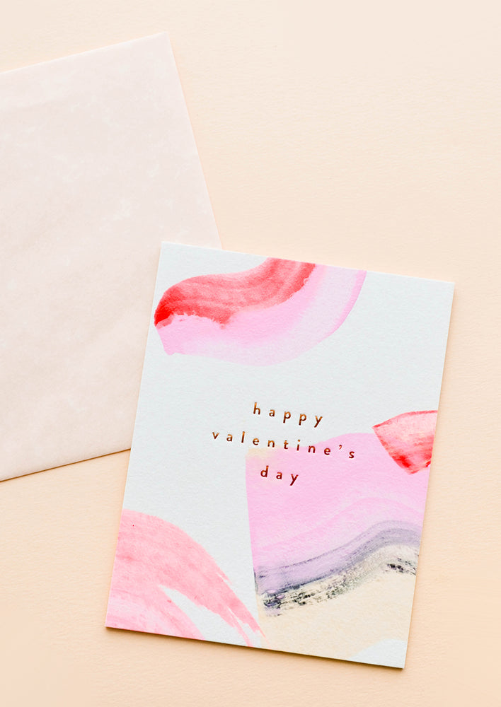 1: A beige envelope and white greeting card patterned with pink and red paint strokes reading "happy valentine's day" in gold foil.
