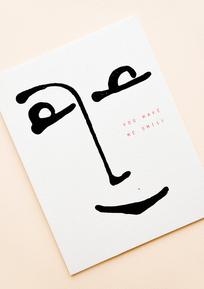 White minimalist greeting card with a simple black drawing of a smiling face and the words "you make me smile" in gold foil.