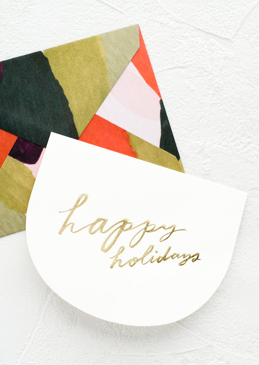 Greeting card in curved shape with gold script reading "Happy holidays", paired with green and red abstract envelope.