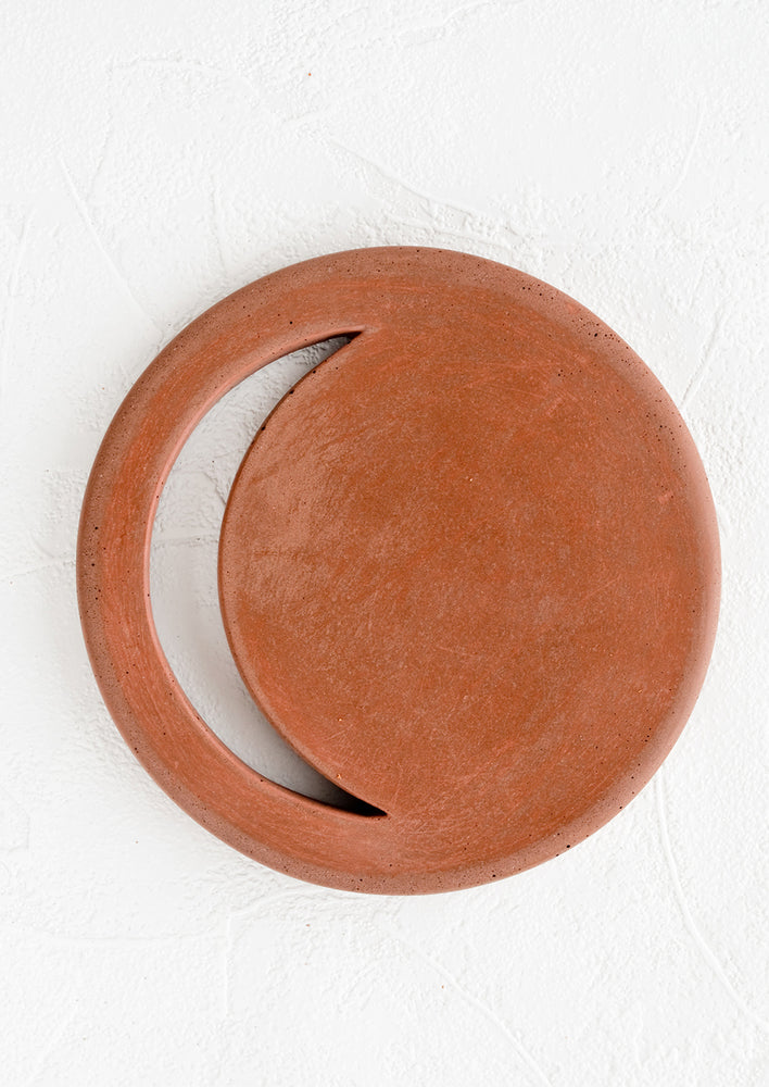 A round concrete trivet in rust color with crescent moon shaped cutout.