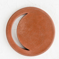 Ore: A round concrete trivet in rust color with crescent moon shaped cutout.