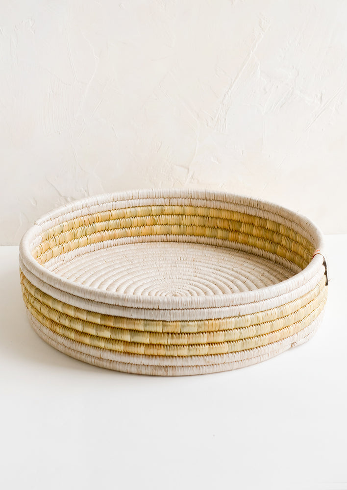 A round, shallow woven seagrass tray in white and beige.