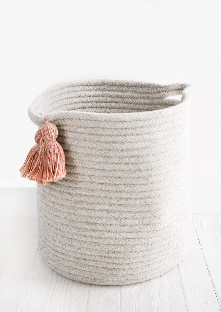 Round, tall storage bin in natural wool. Handles at sides with oversized tassel detail.