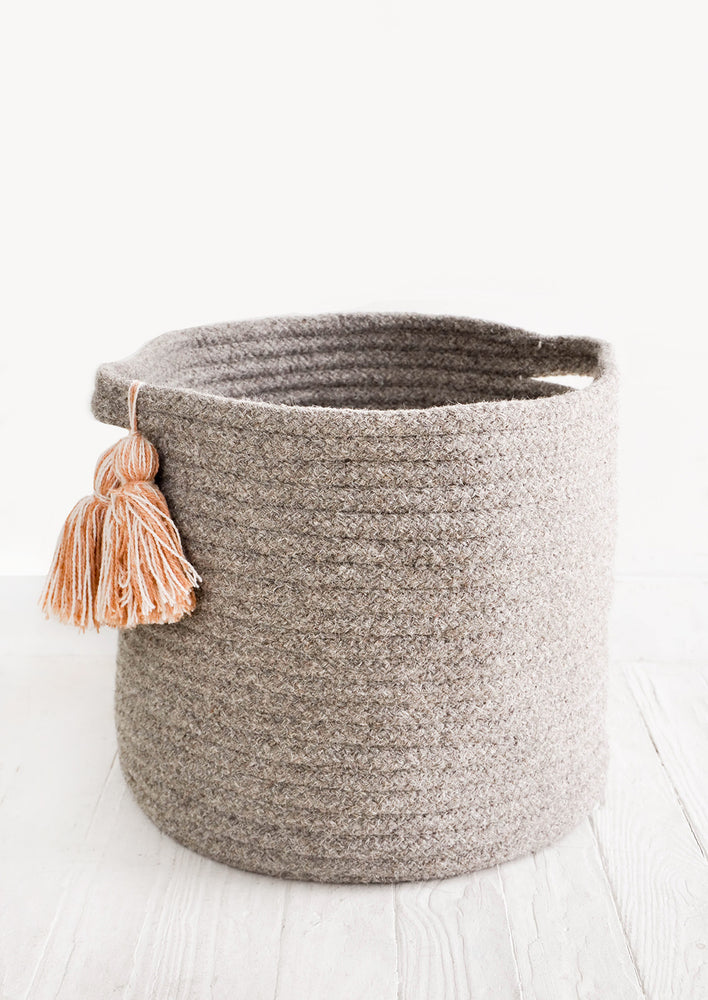 Round, low storage bin in natural wool. Handles at sides with oversized tassel detail.