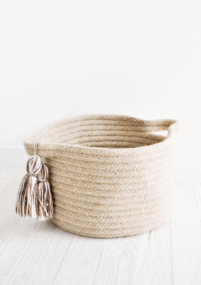 Round, low storage bin in natural wool. Handles at sides with oversized tassel detail.