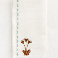 2: A white linen napkin with stitched seafoam border and nile lotus emblem at corner.