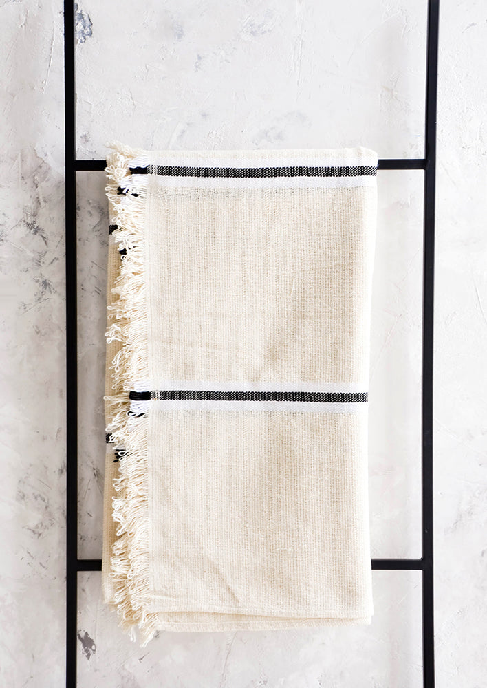 Raw cotton blanket in natural color with black and white stripes and fringed edge, displayed on metal ladder
