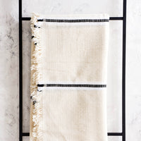 3: Raw cotton blanket in natural color with black and white stripes and fringed edge, displayed on metal ladder