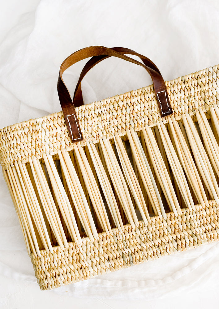 A tote-shaped woven reed basket with open air design, brown leather handles.