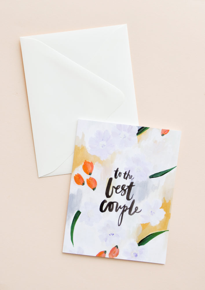 A white envelope and greeting card featuring a painterly floral scene and the words "to the best couple" in black lowercase script.