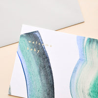 1: A white greeting card with multicolored paint strokes in blue, green, beige, and black and the words "welcome baby" in small lowercase gold foil letters at the top left.