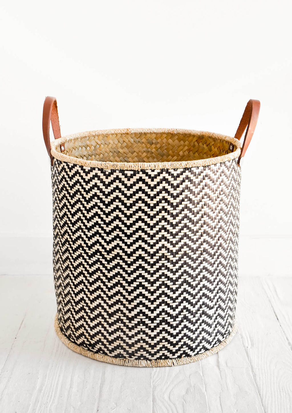 1: Round storage bin in natural material with allover black zigzag pattern and leather handles