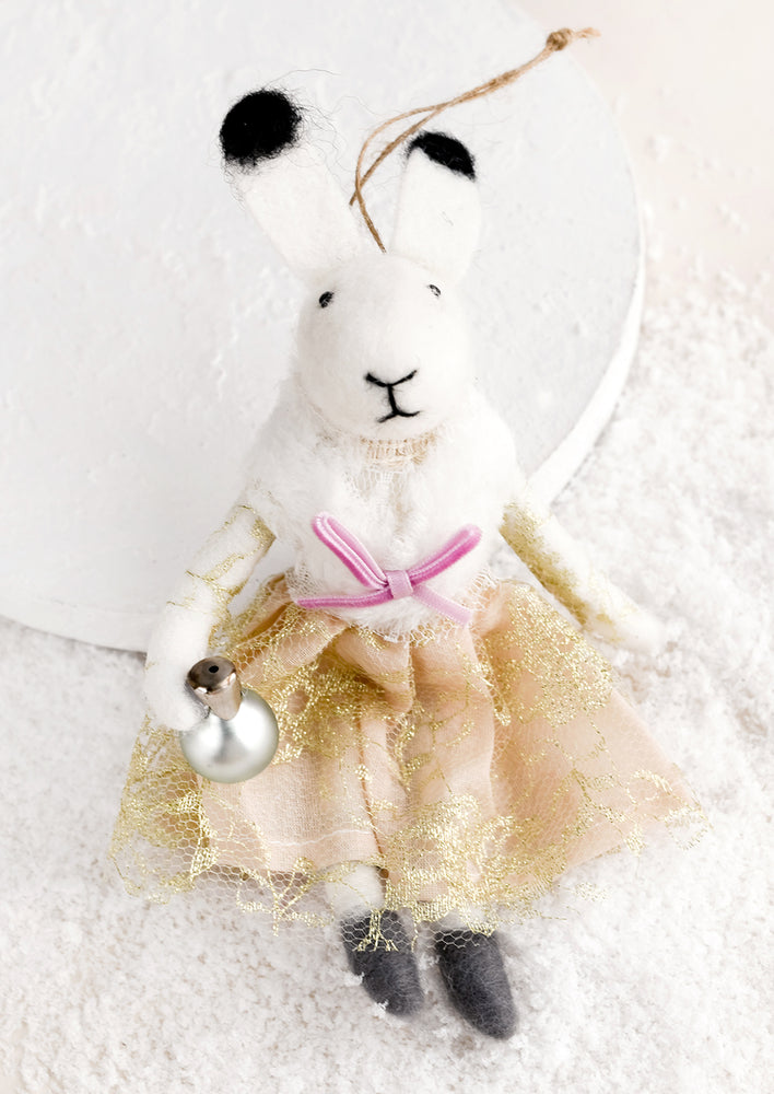 A felted holiday ornament of a bunny wearing a dress, holding a jingle bell.