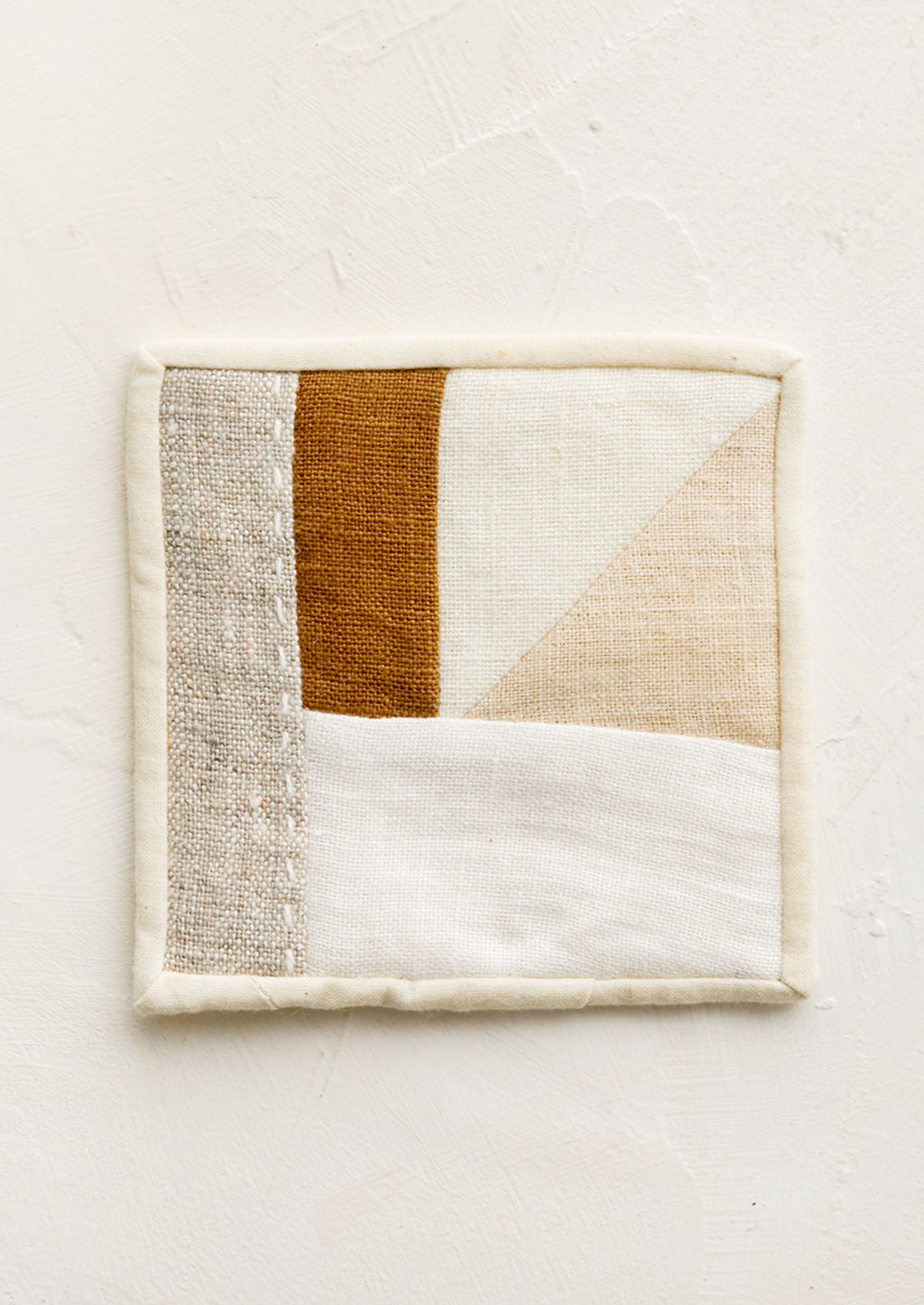 3: A square drink coaster made from neutral hued patchwork fabric.