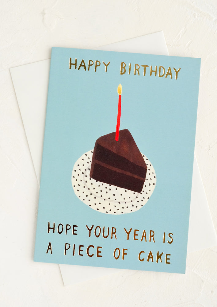A blue greeting card with illustration of chocolate cake and gold text reading "Hope your year is a piece of cake".