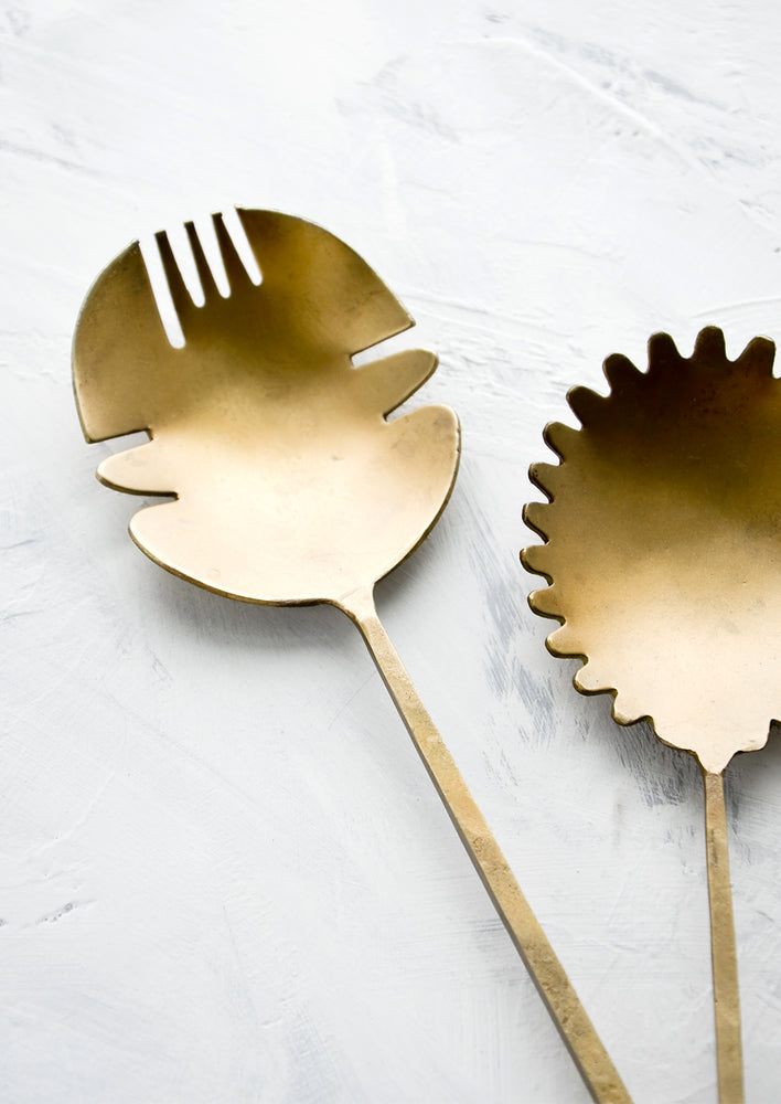 A pair of salad servers made from antiqued brass, shaped in a prehistoric-inspired design showing off cutout and curvy edges.