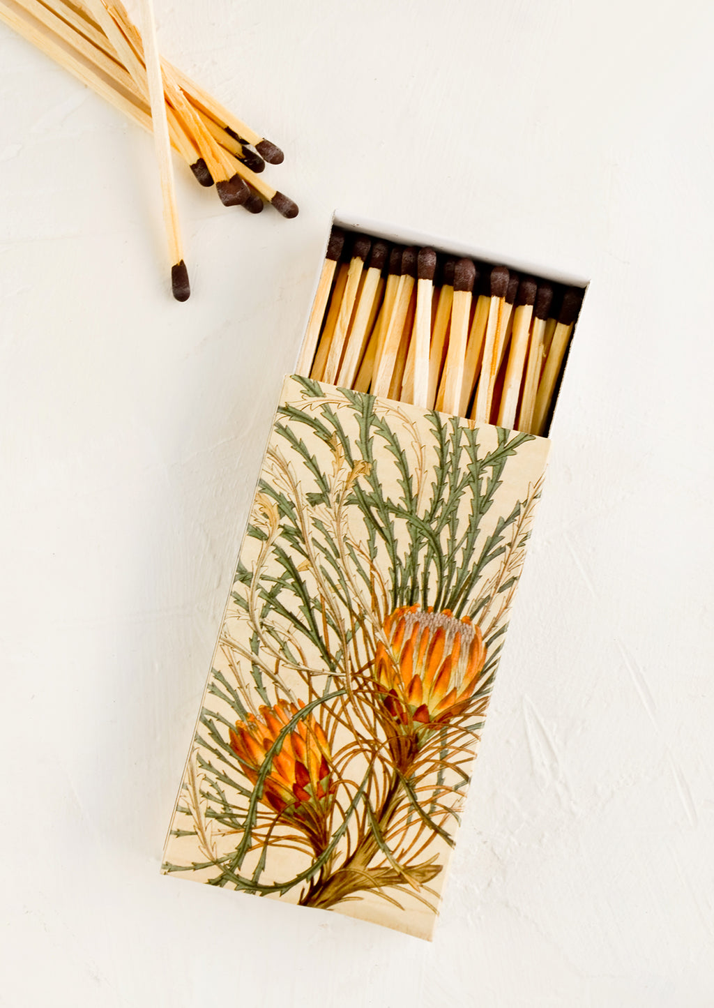 1: A matchbox with botanical protea print and brown-tipped long matches.