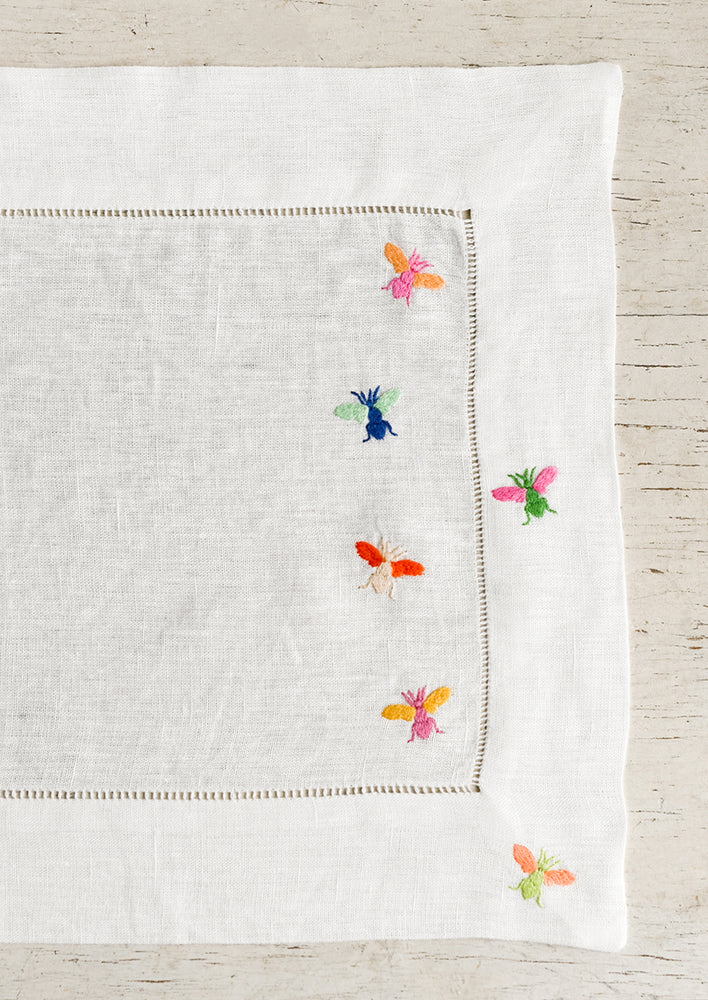 A white linen placemat with multicolor bee embroidery.