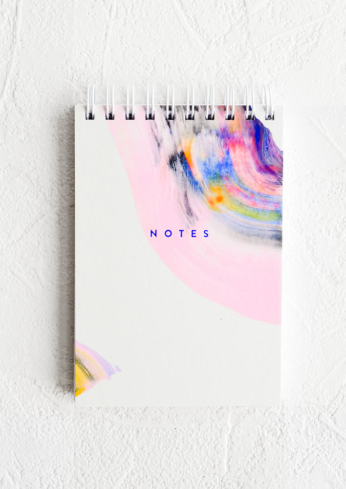A small white spiral bound notepad with rainbow swirl painted cover and "NOTES" on front.