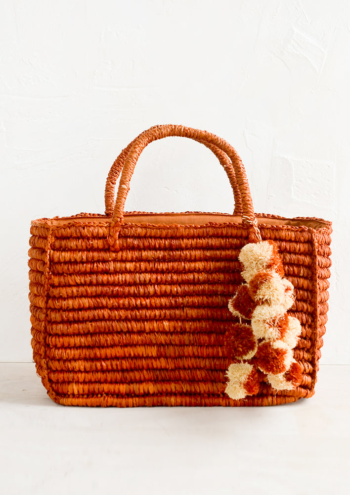 An east-west raffia tote in rust color with straw pom pom detailing.