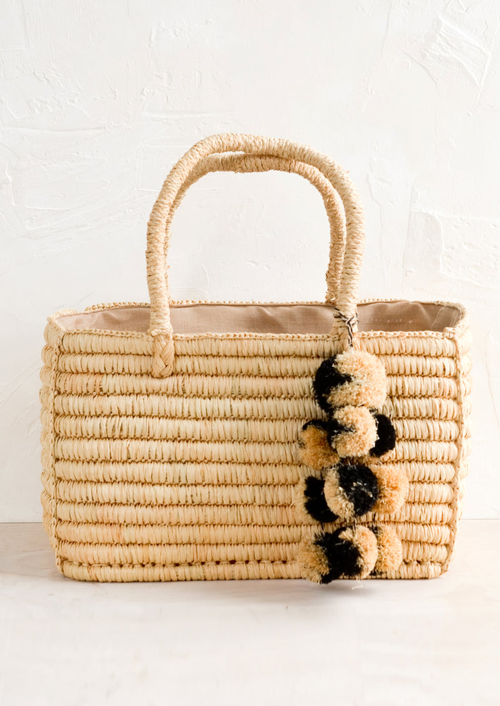 An east-west raffia tote in natural color with black & natural straw pom pom detailing.