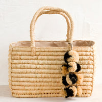 Natural: An east-west raffia tote in natural color with black & natural straw pom pom detailing.