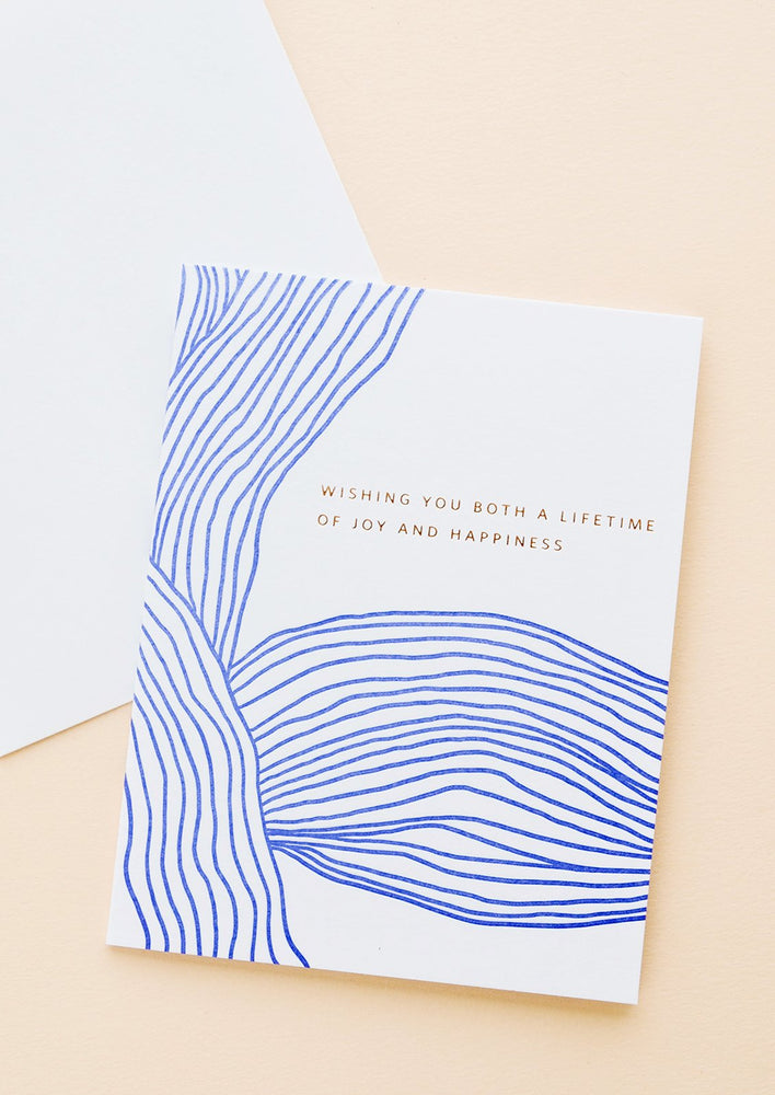 1: White greeting card with wavy cobalt blue line pattern and gold text reading "Wishing you both a lifetime of happiness"