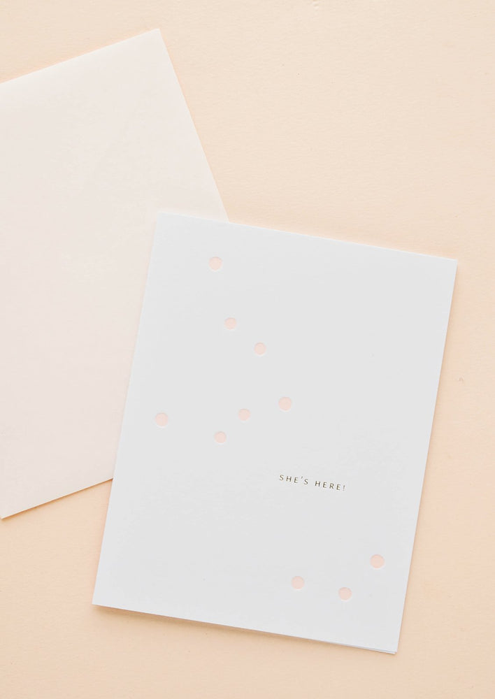 White greeting card with scattered light pink dot and gold text reading "She's here!", paired with pale pink envelope