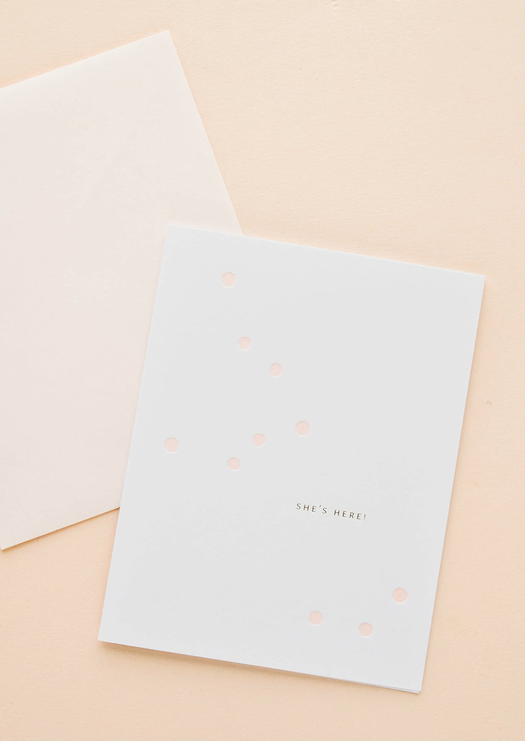 2: White greeting card with scattered light pink dot and gold text reading "She's here!", paired with pale pink envelope