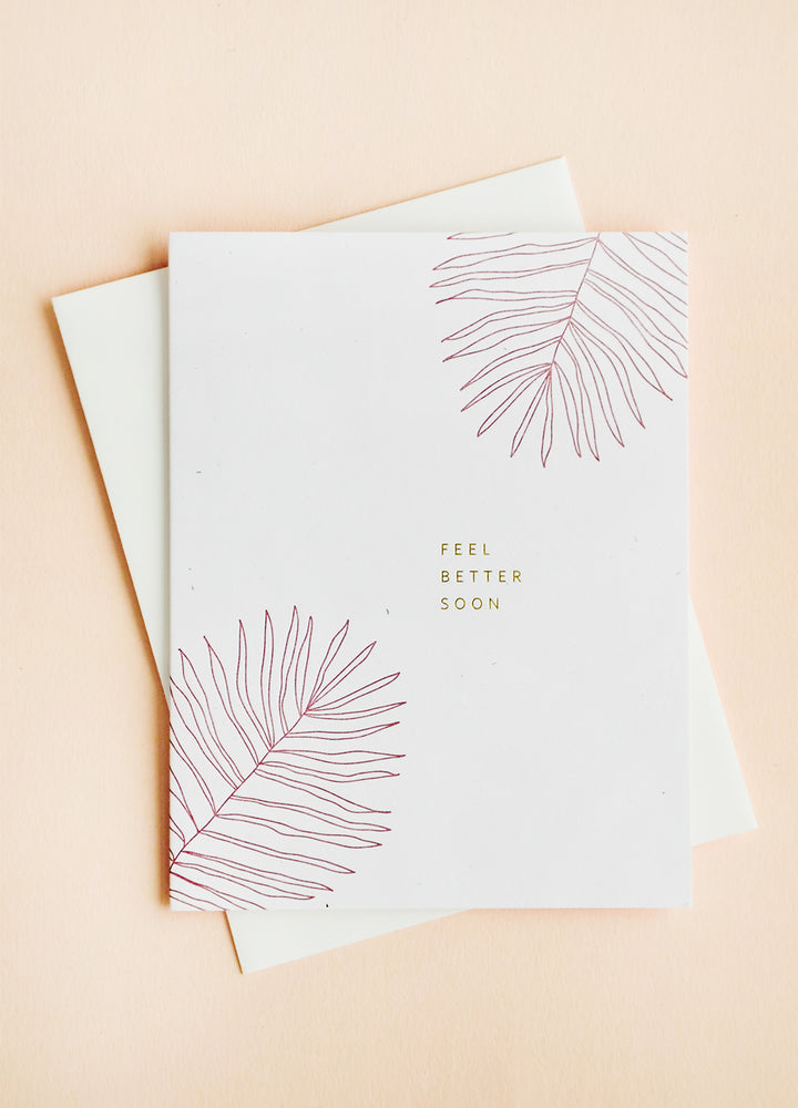 White greeting card and envelope, card features pink palm leaves at corners with small gold text reading "Feel Better Soon".