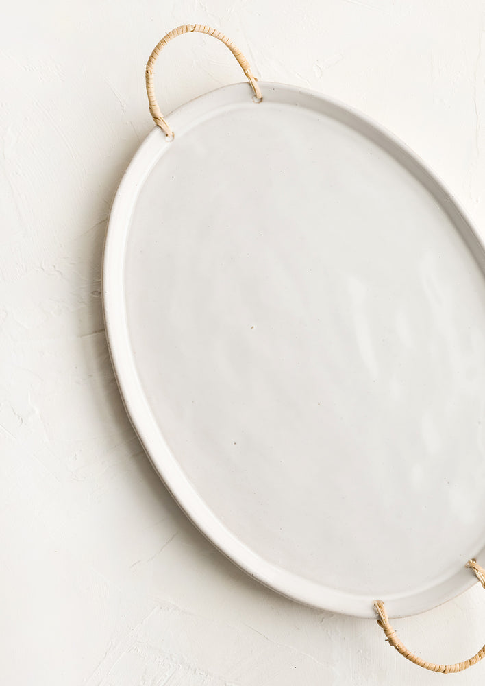 A glossy white ceramic platter in oval shape with rattan wrapped handles at sides.