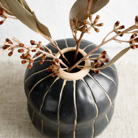 3: A black vase with thin tan vertical stripes, holding dried eucalyptus.