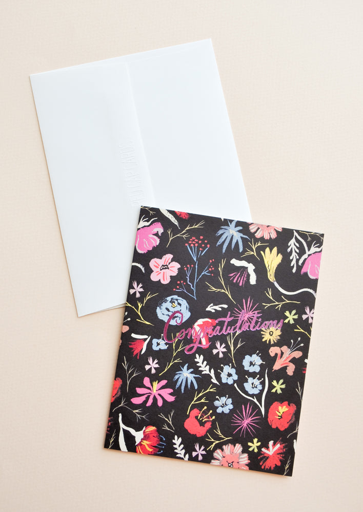Notecard with colorful floral decoration on black background, and the text Congratulations in metallic pink script, with white envelope.